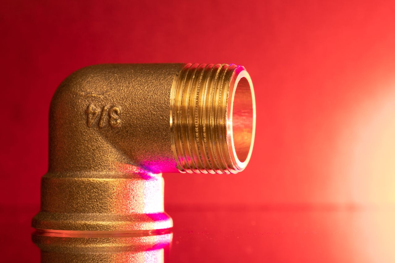our-story-02 Close-up of a shiny brass pipe elbow fitting against a vibrant red backdrop, showcasing industrial elegance.